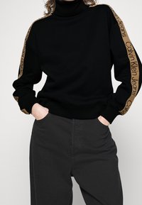Sudadera negra de cuello alto con la marca "Calvin Klein Jeans" en beige en las mangas, combinada con pantalones gris oscuro. Tejido suave, ajuste relajado.