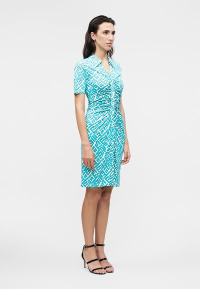 Γυναίκα που φοράει φόρεμα με γεωμετρικό μοτίβο σε τιρκουάζ και λευκό, κοντά μανίκια και ρυτιδωμένο μπροστινό μέρος, με μαύρα σανδάλια με ψηλό τακούνι και λουράκια.