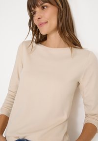 Beige langarmtop met ronde hals, gemaakt van zachte stof. Heeft aansluitende mouwen en een ontspannen pasvorm, minimale naden en geen patronen.