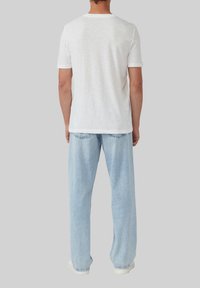 Mann, der mit dem Rücken zur Kamera steht und ein einfaches weißes T-shirt mit kurzen Ärmeln, hellblaue Jeans und weiße Turnschuhe trägt, vor einem grauen Hintergrund.