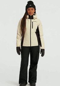 Beige en zwarte geïsoleerde skijas met ritszakken en een vast capuchon, gecombineerd met zwarte skibroek en handschoenen.