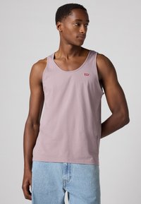 Camisola sem mangas de cor roxa clara, feita de algodão, com um decote redondo e um pequeno logótipo vermelho no peito. Textura suave e design sem mangas.