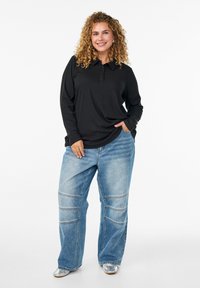 Černé polo tričko s dlouhým rukávem a knoflíkovou légou, v kombinaci se světle modrými kalhotami s širokými nohavicemi, které mají prošité detaily, a stříbrnými botami.
