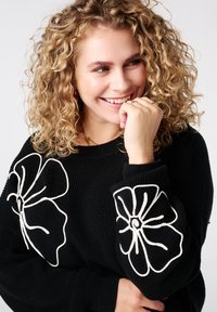 Zwarte trui met witte bloemenborduursels op de mouwen, ribbelstructuur, ronde halslijn en lange mouwen. Model met krullend haar glimlacht.