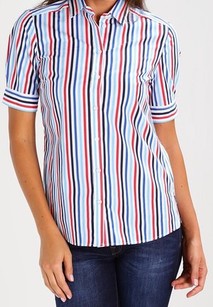 Frau trägt ein kurzärmeliges Hemd mit Kragen mit vertikalen roten, blauen und weißen Streifen, kombiniert mit dunkelblauen Jeans.