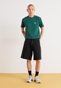 Green short-sleeve t-shirt with a small logo, paired with black denim shorts. Gray athletic shoes with black socks, set against a light backdrop.