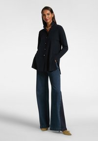 Blusa azul marino con cuello alto, aberturas laterales y botones dorados, combinada con jeans de mezclilla oscuros de pierna ancha y zapatos verde oliva.