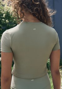 Rövid ujjú, rövid, olívazöld sportfelső, amelyen a hátul egy diszkrét "self." logó található, sima és rugalmas anyagból.