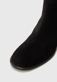 Bottine noire en daim avec un bout rond, texture lisse et une fine semelle en caoutchouc noir. Vue en gros plan montrant les détails du matériau.