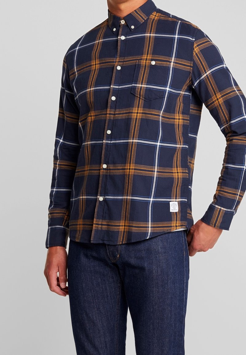 Chemise à carreaux marine à boutons avec motifs orange et bleu, en coton, manches longues et poche poitrine, portée avec un jean denim foncé.
