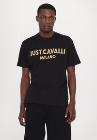 Černé bavlněné tričko s výrazným zlatým grafickým nápisem "JUST CAVALLI MILANO" na přední straně. Krátké rukávy a kulatý výstřih.