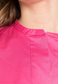 Blusa rosa con scollatura rotonda, texture liscia e riflesso sottile. Presenta un dettaglio cucito che corre lungo la parte anteriore.