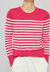 Kobieta w jasnoróżowym i białym poziomo paskowanym swetrze z długimi rękawami, z rękami w kieszeniach białych spodni.