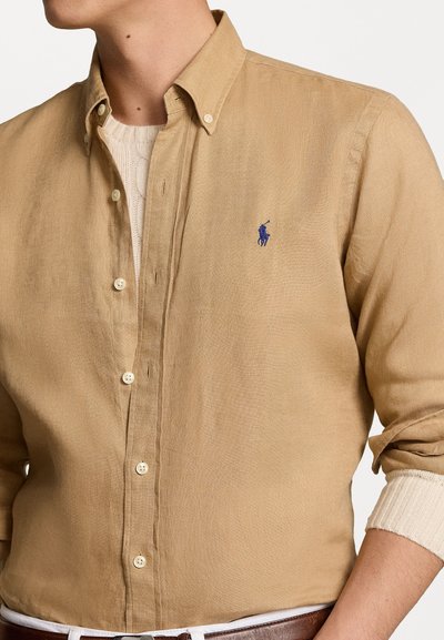 Chemise boutonnée couleur marron clair en lin, dotée d'un col classique et d'un petit logo bleu. Boutons blancs et pull texturé en dessous.