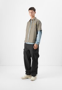Young man stands in a minimalistic setting, wearing a checked short-sleeve shirt over a blue long-sleeve top and black cargo pants.