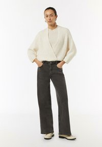 Kremowy kardigan w splot warkocza na lekkim swetrze, zestawiony z szarymi, szerokimi spodniami z wysokim stanem i beżowymi butami. Gładkie tekstury i neutralne kolory.