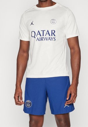 Homme portant le maillot blanc du Paris Saint-Germain avec le logo Qatar Airways et un short bleu avec l'emblème de l'équipe.