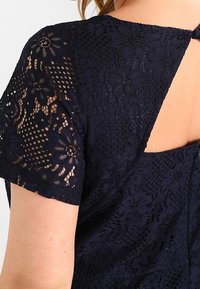 Abito in pizzo blu navy con motivi floreali, maniche corte e dettaglio schiena a goccia chiuso con bottone in tessuto. Design strutturato.