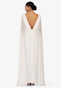 Robe longue blanche avec un décolleté en V plongeant dans le dos, manches longues et fluides, en tissu lisse et léger. Talons visibles au niveau de l'ourlet.