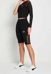 Black long-sleeve crop top with sheer sleeves paired with black bike shorts featuring mesh accents. White sneakers with black and gold details.