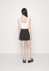 Bílý texturovaný cropped top s volánkovými ramínky, kombinovaný s černými šortkami s vysokým pasem, zobrazený zezadu. Černé platformové boty dotvářejí outfit.