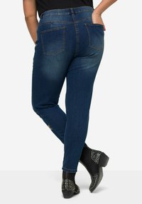 Jeans in denim blu scuro con un design aderente, dotati di due tasche posteriori e una trama liscia. Abbinati a stivali neri con borchie alla caviglia.