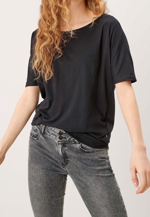 Lockighaarige Person trägt ein locker sitzendes schwarzes Kurzarmshirt und ausgewaschene graue Jeans mit zwei sichtbaren Knöpfen an der Taille.