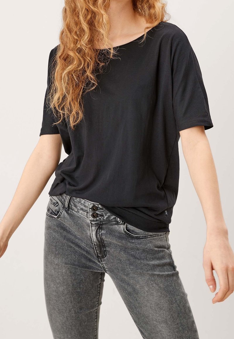 Personne aux cheveux bouclés portant une chemise noire ample à manches courtes et un jean gris délavé avec deux boutons visibles à la taille.