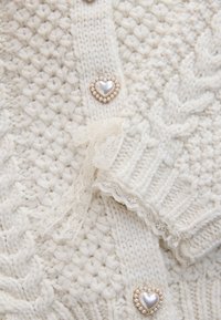 Tissu tricoté crème avec motifs texturés, bordure en dentelle et boutons en forme de cœur bordés de petites perles.