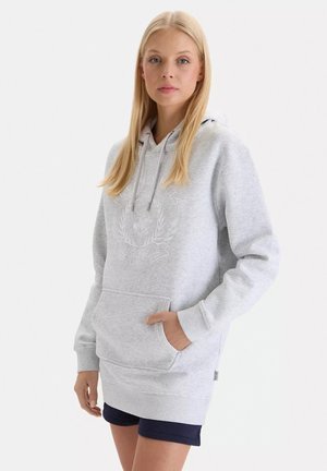 Ung kvinde med langt blondt hår iført en lysegrå hættetrøje og mørke shorts, stående med den ene hånd i frontlommen.
