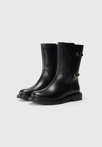 Botas de cuero negro hasta el tobillo con punta redonda, suela plana y hebillas decorativas en el lateral. Textura suave y diseño limpio.