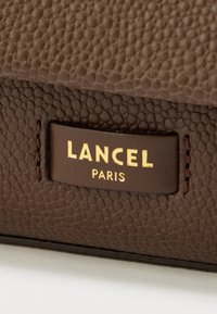 Pruun tekstuuriga nahk, millel on kuldse reljeefse Lanceli logo ja allosas "Paris", millel on peened kividetaolised mustrid.