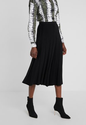Woman wearing a green and white patterned long-sleeve top, black pleated midi skirt, and black pointed-toe ankle boots with thin heels.