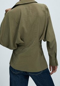 Olivgrön, texturerad blus med en strukturerad krage, ballongsärmar och en åtsittande midja. Matchas med mörk denimjeans.