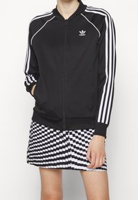 Person som bär en svart Adidas zip-up jacka med vita ränder på ärmarna och en svart-vit rutig kjol.