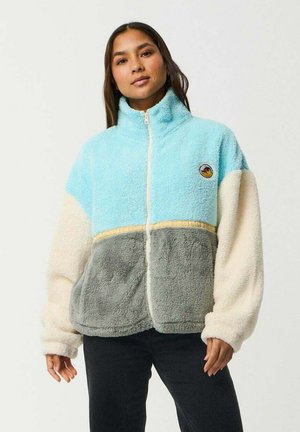 Frau trägt eine Color-Block-Fleecejacke mit hellblauen, cremefarbenen und grauen Abschnitten und steht vor einem schlichten hellen Hintergrund.