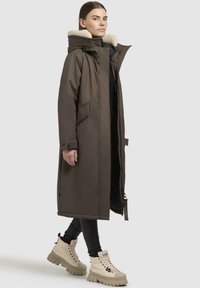 Lang brun parka med en hætte foret med pels, frontlynlås og sidelommer. Båret over sorte leggings og beige tykk-soles støvler. Minimalistisk design.