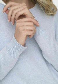 Hellgrauer Pullover mit gerippten Bündchen, der eine glatte Textur und einen lässigen Schnitt aufweist. Sichtbare Hände zeigen natürliche Nägel und einen entspannten Griff.