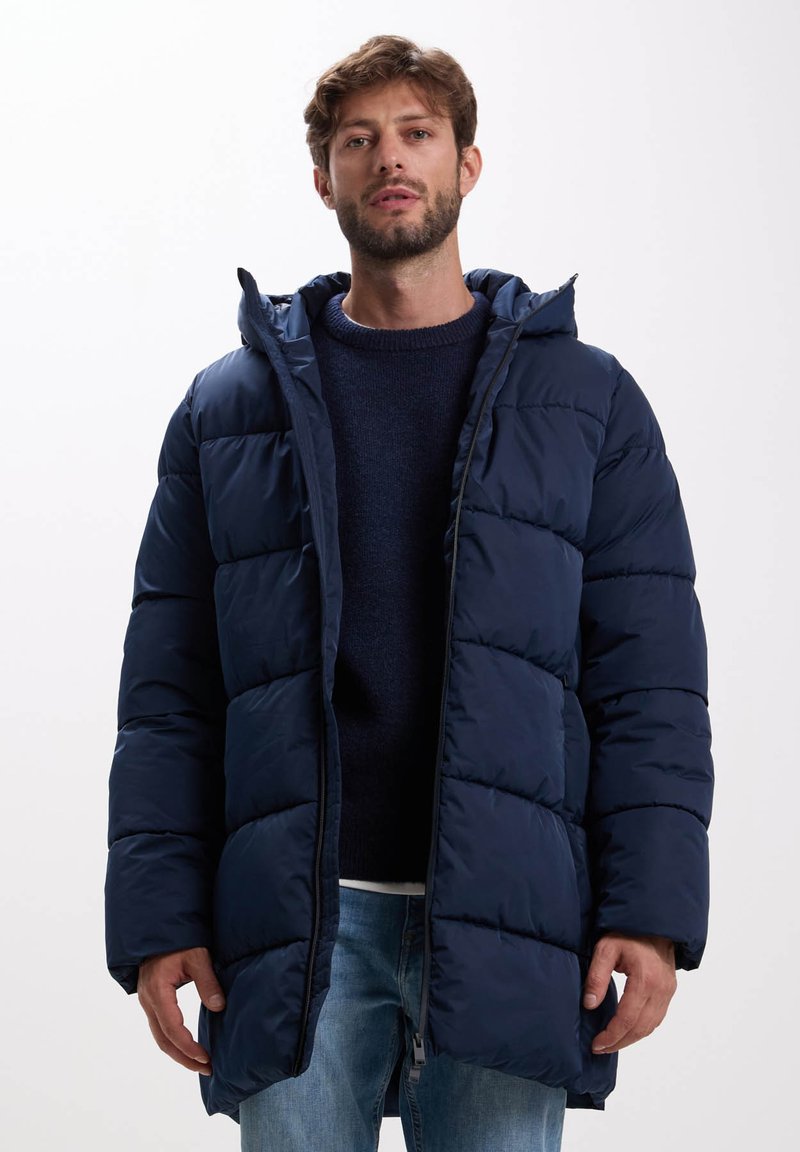Manteau en duvet bleu marine avec capuche, présentant un design matelassé, une fermeture éclair à l'avant et des poches latérales. Porté par-dessus un pull sombre et un jean clair.