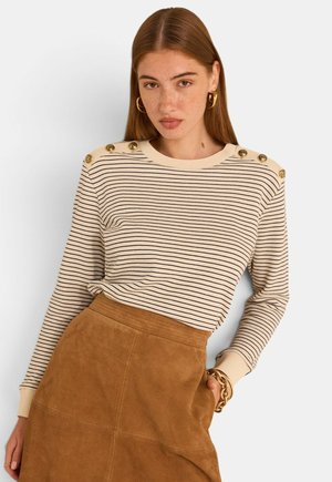 Jeune femme aux longs cheveux bruns portant un pull beige et noir à rayures avec des boutons aux épaules et une jupe en suède marron, debout contre un mur blanc.
