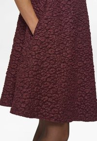 Burgundy színű ruha, amelynek texturált felülete apró, kiemelkedő virágmintákkal díszített. Oldalsó zsebekkel és bővülő szegéllyel rendelkezik.