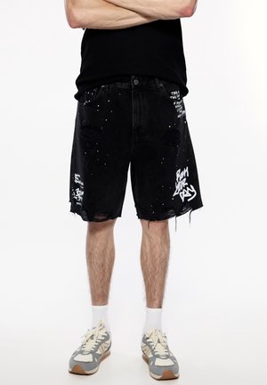 Homme portant un short en jean noir usé avec un texte graffiti blanc et des éclaboussures de peinture, associé à des chaussettes blanches et des baskets grises.
