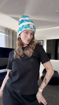 Juodas trumpomis rankovėmis marškinėlis derintas su tamsiomis kelnėmis. Mėlynas ir baltas raštuotas megztas kepuraitė. Lygūs plaukai, auksinės auskarai ir matomi tatuiruotės.