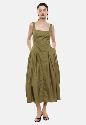 Donna che indossa un vestito midi verde oliva senza maniche con scollatura quadrata e gonna plissettata, abbinato a tacchi aperti scuri e orecchini pendenti.