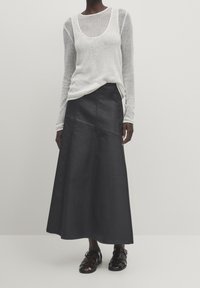 Gonna lunga in denim nero con taglio dritto e cuciture ben visibili, abbinata a una blusa bianca leggera e trasparente con maniche lunghe.