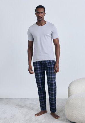 Mand stående barfodet iført lysegrå kortærmet T-shirt med "Hugo"-logo og marineblå ternede pyjamasbukser på lysegråt tæppe.