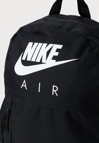 Musta reppu, jossa on teksturoitu kangas, varustettuna suurella valkoisella "NIKE"-logolla ja "AIR"-tekstillä. Sisältää etutaskun vetoketjulla ja kaksi yläkahvaa.
