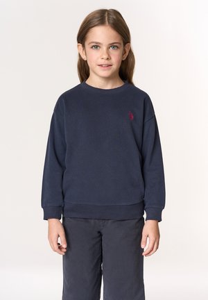 Felpa blu navy in tessuto morbido con polsini e orlo a coste, caratterizzata da un piccolo logo rosso ricamato sul petto a sinistra. Indossata con pantaloni scuri.