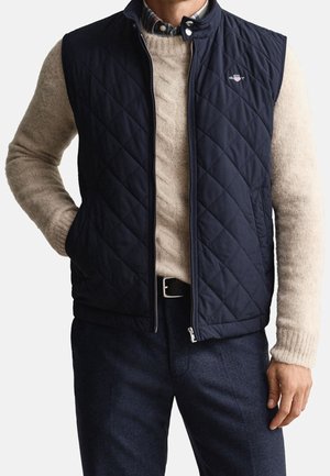 Uomo che indossa un gilet trapuntato blu navy sopra un maglione beige a trecce e pantaloni scuri, in piedi con una mano in tasca.