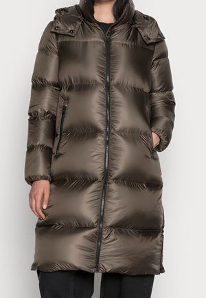 Kobieta ubrana w długi, błyszczący, brązowy pikowany płaszcz z frontowym suwakiem i bocznymi kieszeniami na jednolitym jasnym tle.